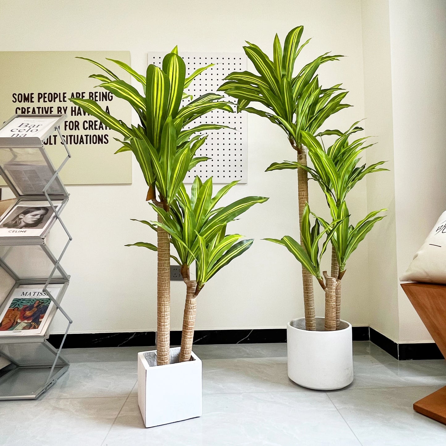 Deux plantes artificielles d'intérieur (Dracaena) aux feuilles vertes et rayées apportent de la fraîcheur à un espace de vie moderne. Sur le carrelage clair, elles sont disposées près d'un porte-revues, d'une décoration murale avec texte et d'une partie d'un fauteuil rembourré en arrière-plan.