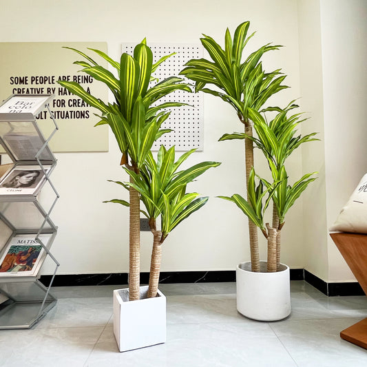 Deux plantes artificielles d'intérieur (Dracaena) aux feuilles vertes et rayées apportent de la fraîcheur à un espace de vie moderne. Sur le carrelage clair, elles sont disposées près d'un porte-revues, d'une décoration murale avec texte et d'une partie d'un fauteuil rembourré en arrière-plan.