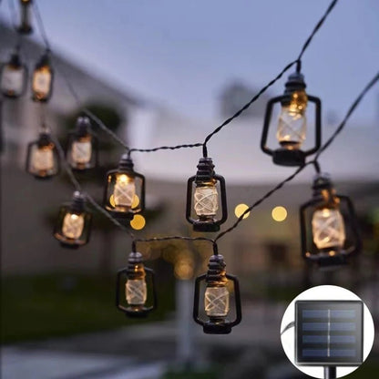A la tombée de la nuit, les guirlandes lumineuses GUIRLANDE EXTERIUR SOLAIRE | Lumière Heureuse en forme de lanterne s'illuminent d'une lumière chaleureuse rappelant Noël. Le fond flou évoque un décor de jardin. Un encart confirme qu'il s'agit de lampes solaires LED équipées d'un panneau solaire.