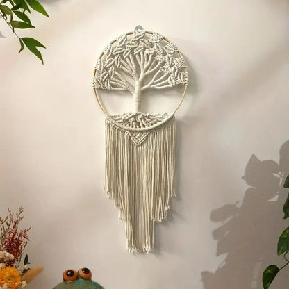 Un arbre de vie en macramé crémeux orne un mur blanc, suspendu à un cerceau de bois clair. Les détails complexes des nœuds forment un tronc noueux et des branches délicates. De longues franges pendantes ajoutent une texture douce à cette décoration murale artisanale.