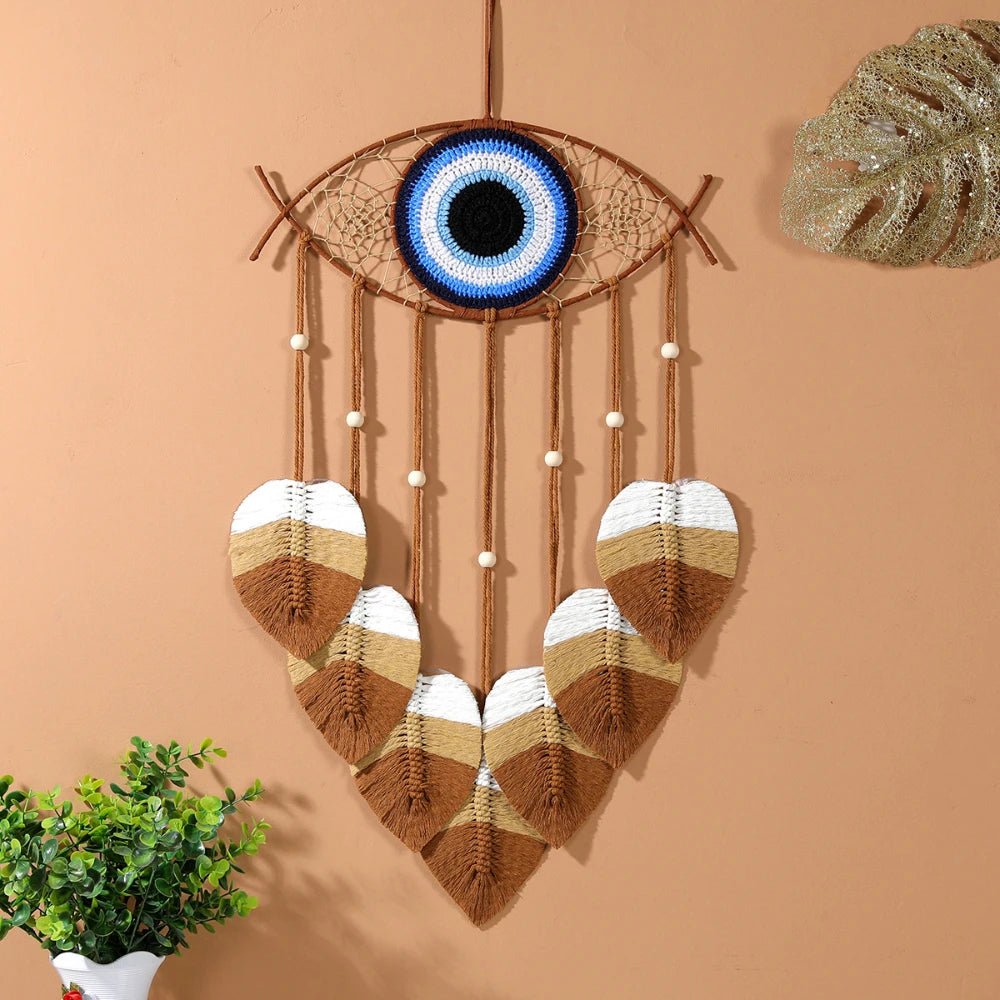 Un attrape-rêves en macramé unique orné d'un œil bleu protecteur au centre. Des plumes décoratives en macramé dans des tons naturels de blanc, beige et marron pen macramé unique orné d'un œil bleu protecteur au centre. Des plumes décoratives en macramé dans des tons naturels de blanc, beige et marron pendent délicatement. L'ensemble est suspendu sur un mur de couleur pêche, créant une ambiance bohème et spirituelle. Une petite plante verte en pot ajoute une touche de fraîcheur dans le coin inférieur gauche.