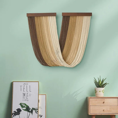Un élégant ouvrage en macramé suspendu au mur vert pâle, fait de cordes beiges et marron. La pièce de macramé a une forme fluide et ondulée, fixée par deux barres en bois sombre. En dessous, on aperçoit un coin de pièce avec une plante et un cadre décoratif. La texture délicate du macramé contraste avec la simplicité du mur.
