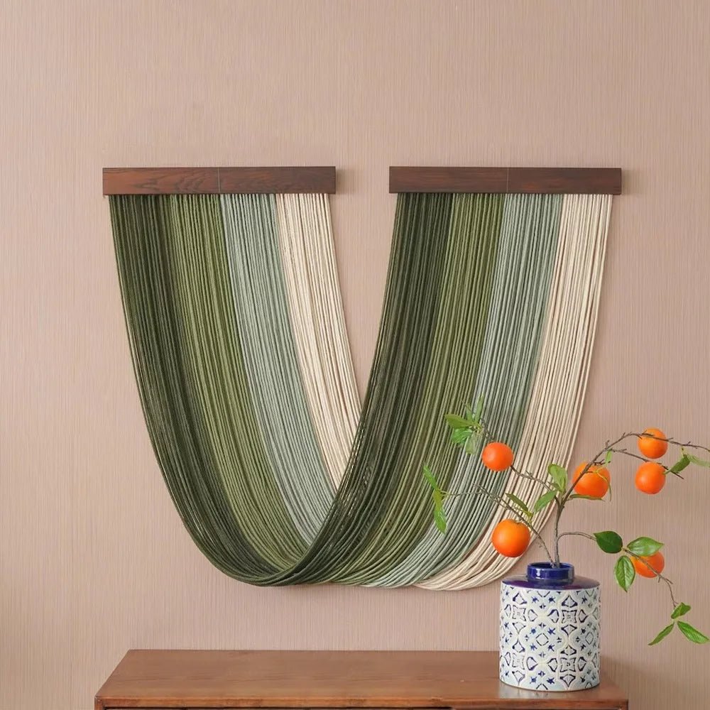 Un élégant panneau décoratif en macramé, suspendu à deux barres de bois foncé, orne un mur beige texturé. Les cordes, dans des nuances de vert et de crème, forment un motif fluide et captivant. Sur une commode en bois, un petit vase bleu et blanc soutient une branche de kaki aux fruits orange vif, ajoutant une touche de nature à la scène.