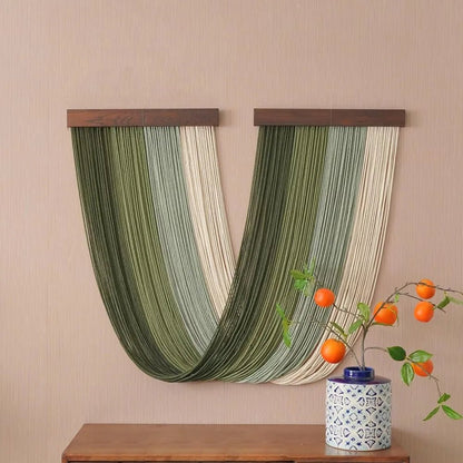 Un élégant panneau décoratif en macramé, suspendu à deux barres de bois foncé, orne un mur beige texturé. Les cordes, dans des nuances de vert et de crème, forment un motif fluide et captivant. Sur une commode en bois, un petit vase bleu et blanc soutient une branche de kaki aux fruits orange vif, ajoutant une touche de nature à la scène.