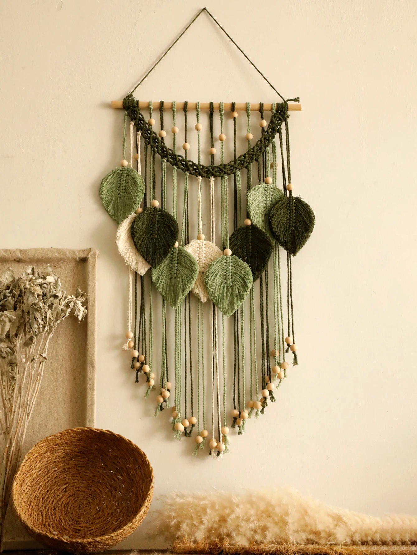 Un macramé décoratif suspendu à un mur beige clair présente un motif de feuilles vertes et beiges. Des perles de bois clair parsèment les cordes et ornent le bas de la pièce. L'art mural crée une ambiance bohème et naturelle. Marcame.