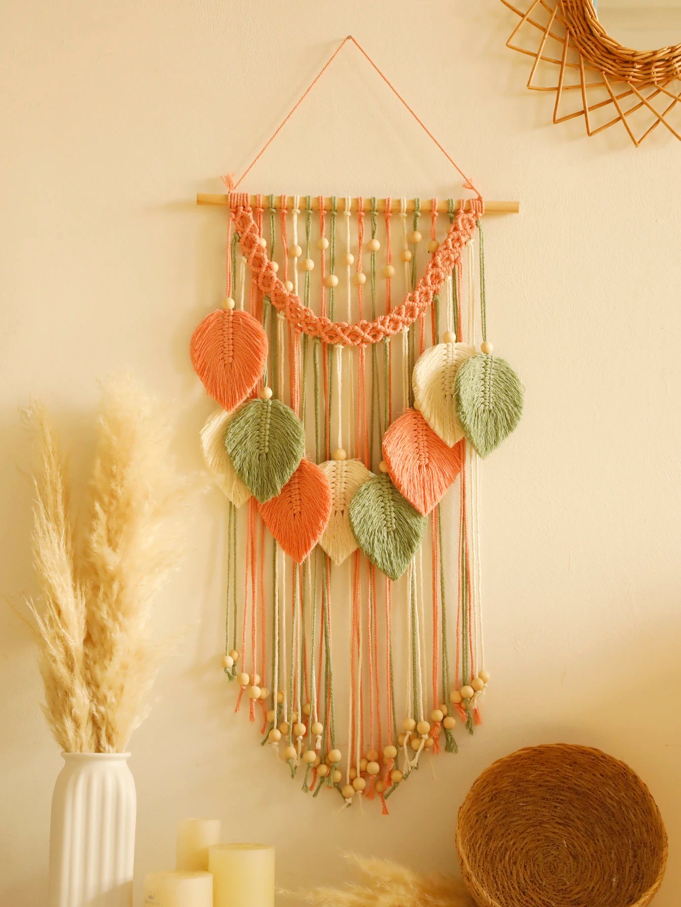 Un magnifique macramé décoratif orne un mur crème, suspendu à une baguette en bois. Des feuilles en macramé de couleurs pêche, vert sauge et crème sont délicatement nouées, créant un motif bohème. Des perles de bois parsèment les cordes et la bordure supérieure. Marcrame artisanal apportant une touche chaleureuse à l'intérieur.