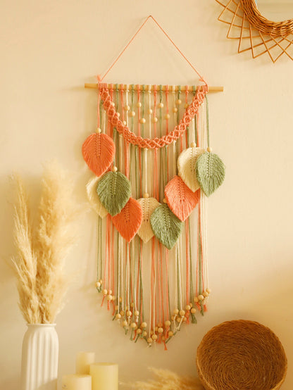 Un magnifique macramé décoratif orne un mur crème, suspendu à une baguette en bois. Des feuilles en macramé de couleurs pêche, vert sauge et crème sont délicatement nouées, créant un motif bohème. Des perles de bois parsèment les cordes et la bordure supérieure. Marcrame artisanal apportant une touche chaleureuse à l'intérieur.