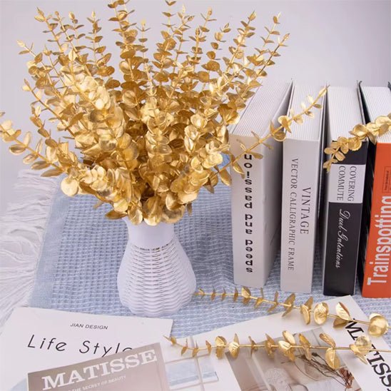 Un vase blanc avec "Plantes artificielles comme des vraies-Feuilles D'eucalyptus Doré" se dresse sur une table à côté de livres debout et d'un magazine "Life Style", créant une ambiance de décoration d'intérieur moderne.