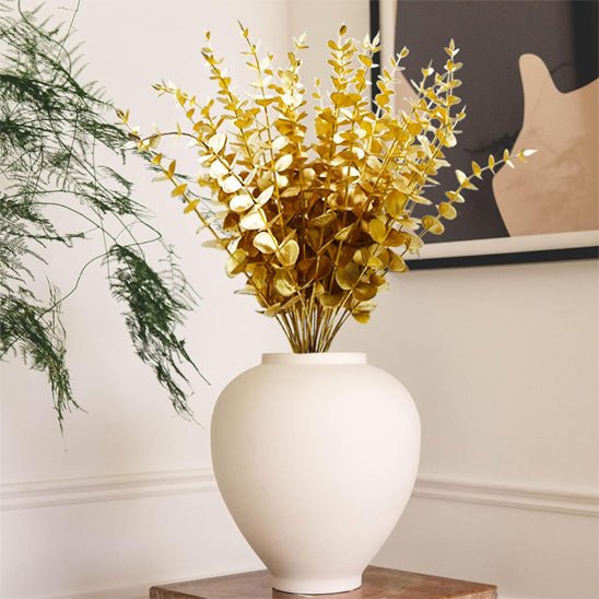 Un vase en céramique blanche orné de Plantes artificielles comme des vraies – Feuilles d'eucalyptus dorées – est posé sur une table en bois, mettant en valeur le décor. Une plante à feuilles vertes se trouve à gauche, et une œuvre d'art abstrait décore le mur blanc en arrière-plan.