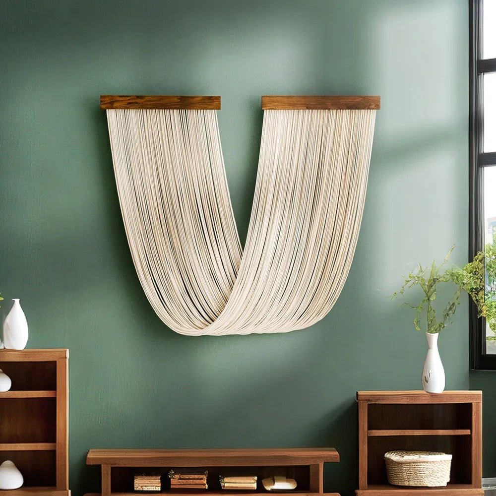 Une décoration murale en macramé beige clair est suspendue à deux supports en bois foncé sur un mur vert olive. Les cordes forment une courbe élégante, apportant une touche artisanale et bohème à l'espace. La texture du macramé crée un contraste subtil avec la surface lisse du mur. Cette pièce de macramé unique est un élément de décoration intérieure artistique.