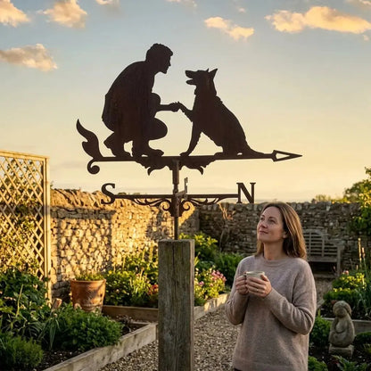 Une girouette animal en fer forgé représentant la silhouette d'une personne et d'un chien sous un ciel bleu nuageux.