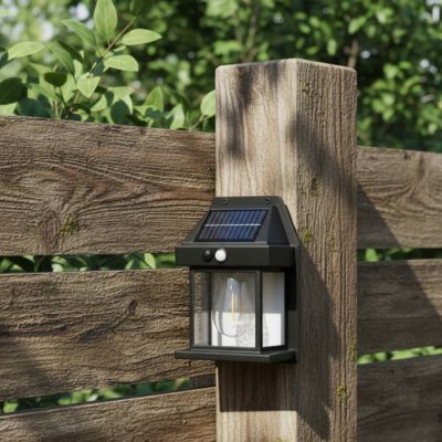 Une lampe solaire jardin haut de gamme de couleur noire fixée sur un pilier en bois au milieu d'un espace extérieur verdoyant en journée.