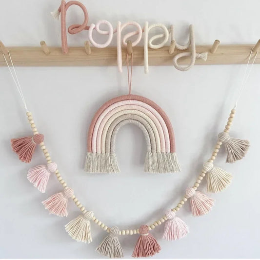 Décoration murale de chambre d'enfant présentant le prénom "Poppy" en fil rose clair et blanc cassé, un arc-en-ciel suspendu en macramé dans des tons de rose poudré, beige et blanc, ainsi qu'une guirlande de perles de bois et de pompons en macramé assortis. L'ensemble est accroché à un porte-manteau en bois clair sur un mur blanc. Cette décoration douce et artisanale crée une ambiance chaleureuse et personnalisée.