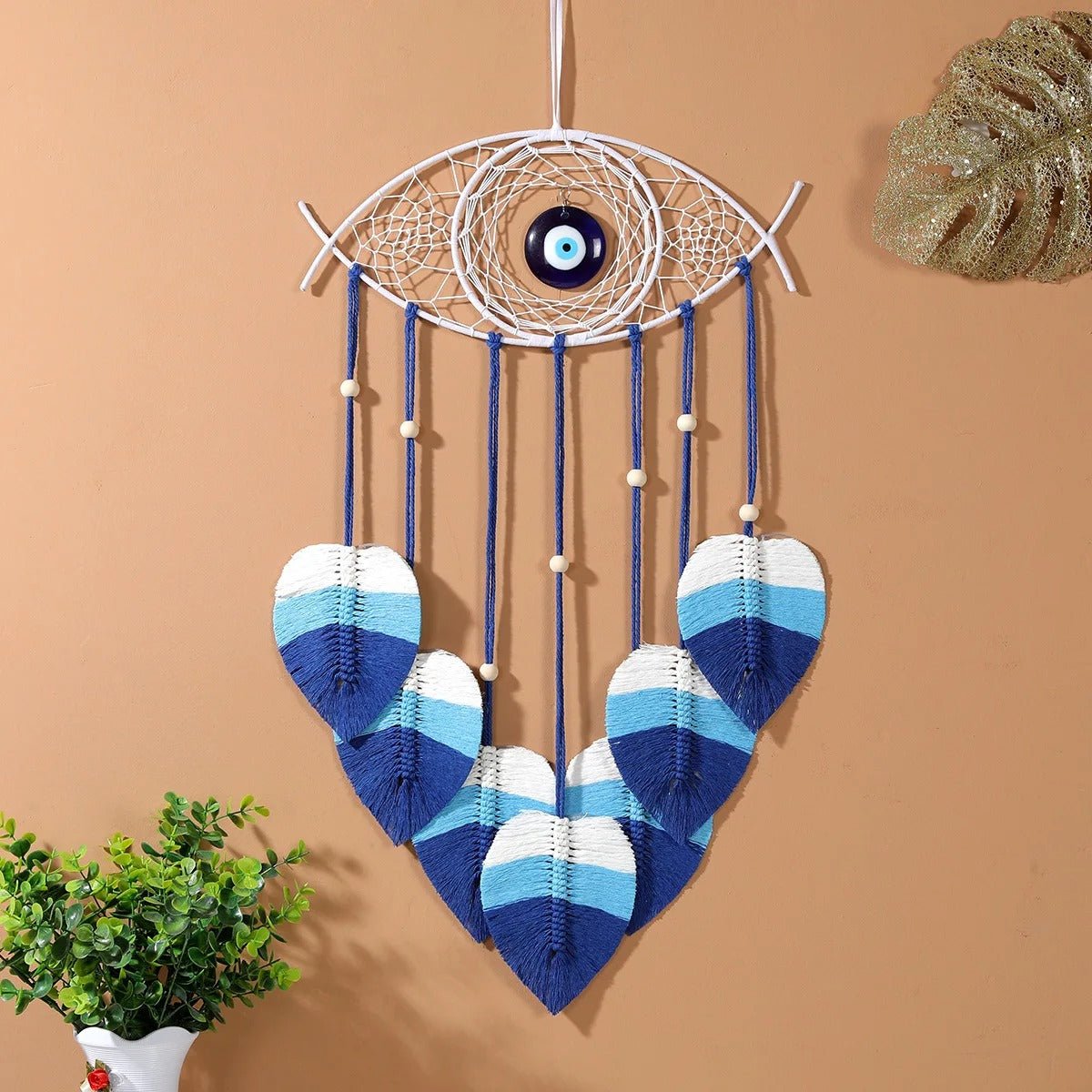 Un attrape-rêves décoratif en macramé blanc en forme d'œil avec un œil bleu au centre est suspendu à un mur pêche. De fines cordes bleues ornées de perles de bois claires pendent du bas, chacune se terminant par une feuille en macramé dégradée de blanc, bleu clair et bleu foncé. Une petite plante verte en pot blanc est visible en bas à gauche et une feuille décorative dorée est partiellement visible en haut à droite.