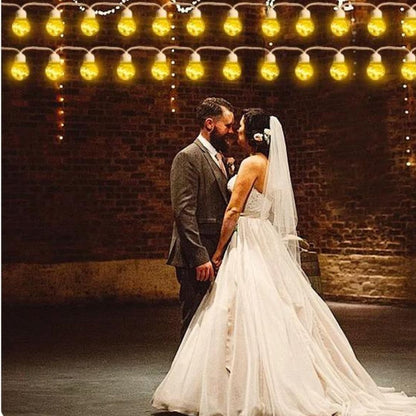 Une mariée dans une robe blanche fluide avec un voile et le marié en costume se tiennent côte à côte dans un espace faiblement éclairé sous une voûte enchanteresse de guirlandes solaires extérieures EcoLume, qui projettent des lueurs chaudes sur le mur de briques rustique.