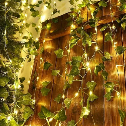Des guirlandes lumineuses festives mélangées à des feuilles de lierre vertes et "Guirlande solaire extérieure | Lueur de bonheur" sont accrochées sur un fond de bois, créant une ambiance féerique. La douce lueur jaune enrichit la texture et les feuilles du bois, offrant des options de décoration polyvalentes.