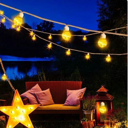 Un coin salon extérieur cosy est composé dun canapé en osier avec coussins, éclairé par des guirlandes lumineuses et des guirlandes solaires extérieures EcoLume. Un lampadaire en forme détoile et une lanterne rehaussent lambiance, le tout conçu pour une protection optimale IP44.