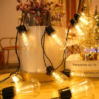 La guirlande solaire SolarZen aux ampoules lumineuses est drapée autour dun pot blanc orné de fleurs séchées, créant une ambiance agréable. La lueur chaleureuse renforce latmosphère chaleureuse, tandis quune pile de livres et une décoration en spirale dorée trônent en arrière-plan.