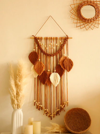  décoration murale en macramé aux tons chauds, suspendue sur un mur beige. L'ouvrage artisanal présente des motifs de feuilles en différentes nuances de brun et de blanc cassé, agrémentées de perles en bois. Un miroir en rotin en forme de soleil et un panier tressé complètent le décor bohème et naturel. Un vase blanc contenant des herbes de la pampa est visible sur une surface en dessous. Marcame.