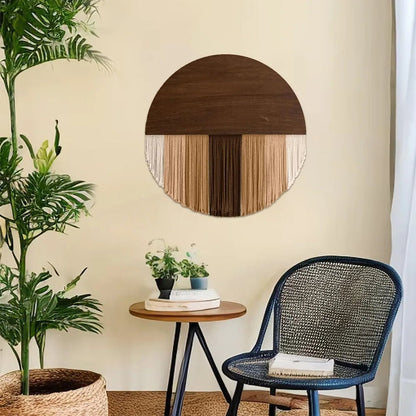 Image d'une décoration murale en macramé suspendue au-dessus d'une petite table ronde en bois avec des plantes. La pièce maîtresse circulaire combine un demi-disque en bois foncé avec des franges de cordes en coton de couleurs naturelles, créant un effet bohème chic. Un fauteuil en rotin noir et une plante verte en pot complètent le décor intérieur minimaliste et chaleureux.