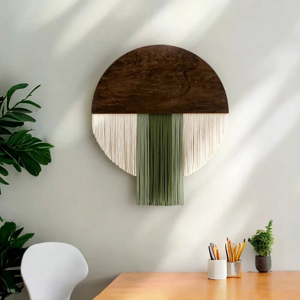 Une décoration murale en macramé unique et élégante, présentant un disque en bois sombre surmonté de franges de corde beige et verte tombant en cascade. L'œuvre d'art crée une ambiance bohème et artisanale, parfaite pour ajouter une touche de texture et de chaleur à n'importe quelle pièce. Les détails complexes du macramé et le contraste des couleurs en font une pièce maîtresse captivante.