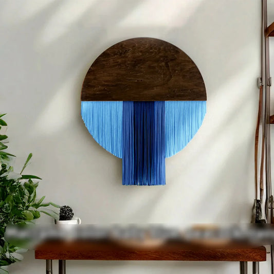 Une décoration murale unique en macramé, présentant un demi-cercle en bois sombre surmonté de franges de fils bleu clair et bleu foncé. L'œuvre d'art apporte une touche bohème et artisanale à tout intérieur. Elle est suspendue sur un mur blanc, créant un contraste visuel attrayant.