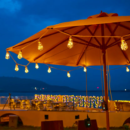 Un parasol de terrasse avec des guirlandes lumineuses illumine la soirée, tandis que les guirlandes solaires extérieures de SunGlow ornent une balustrade, créant une ambiance chaleureuse sur fond de ciel crépusculaire et de montagne.
