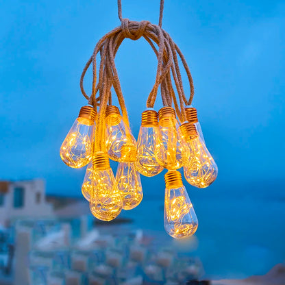 Les guirlandes solaires extérieures SunGlow, en forme d'ampoules, sont suspendues à une ficelle épaisse sur fond de vue floue sur la côte. Le ciel couvert et la mer visible renforcent l'ambiance chaleureuse.
