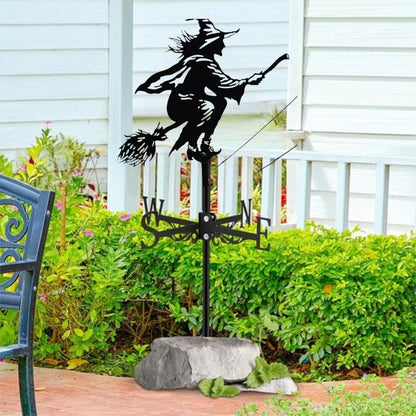 La girouette de toit-sorcière, ornée d'une silhouette de sorcière sur un balai, est fixée sur un rocher de jardin, ajoutant une touche de magie au décor. Ornée des lettres directionnelles N, E, S et O, elle se dresse avec charme sur fond de bâtiment blanc et de buissons verdoyants.