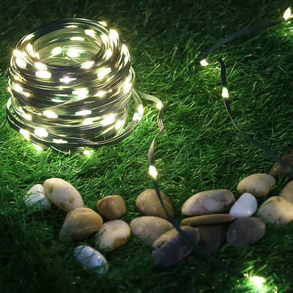 Une guirlande lumineuse solaire extérieure SolarSpark est placée sur l'herbe verte, illuminant les alentours. Des pierres lisses et multicolores se dispersent près des lumières, renforçant l'atmosphère sereine et décorative de l'extérieur.