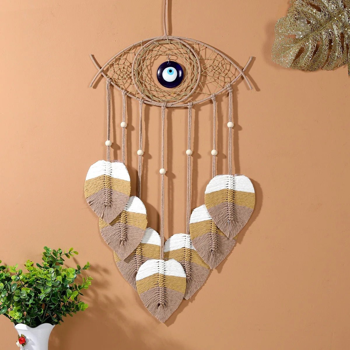 Un attrape-rêves en macramé en forme d'œil orné d'une perle bleue au centre, suspendu à un mur pêche. Des cordons en macramé beige pendent, agrémentés de perles blanches et terminés par des feuilles décoratives en macramé aux couleurs blanc, beige et marron. Une petite plante verte en pot est visible en bas à gauche.