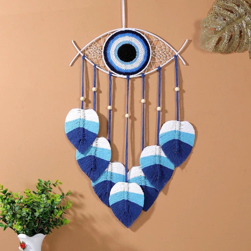 Un capteur de rêves en macramé en forme d'œil bleu et blanc est suspendu à un mur pêche. Des cordes bleues ornées de perles de bois pendent, terminées par des feuilles en macramé bleu et blanc dégradé. Une petite plante verte en pot blanc est visible en bas à gauche. Un ornement doré en forme de feuille est fixé sur le mur à droite.