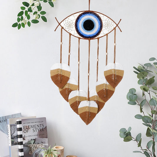 une décoration murale en macramé en forme d'œil.
L'œil en macramé présente un centre noir, entouré de bleu et de blanc.
Des cordes marron pendent de l'œil, avec des feuilles décoratives en macramé blanc, beige et marron.