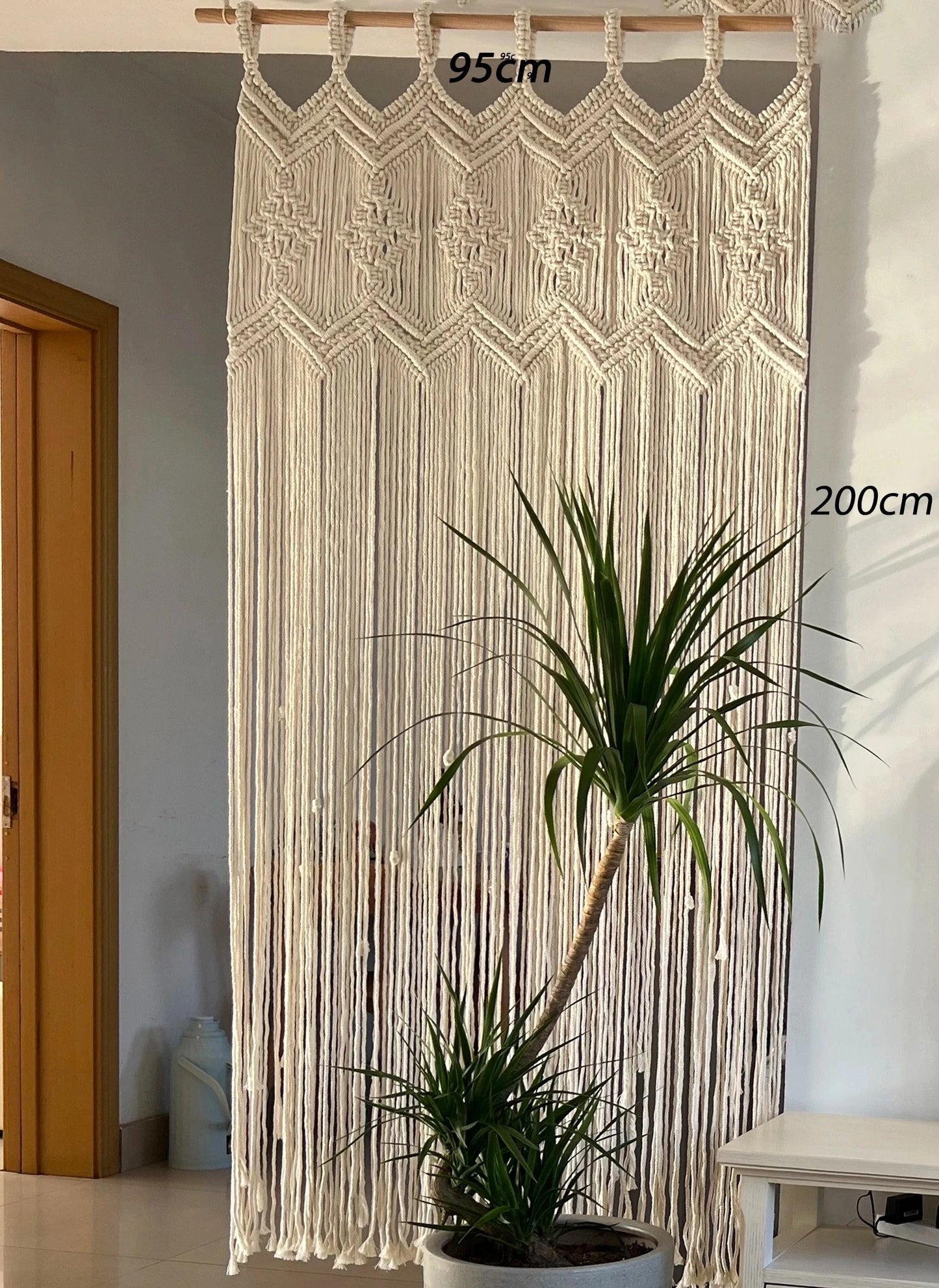 Un  macramé rideau crème, mesurant 95 cm de largeur et 200 cm de hauteur, orne le mur à côté d'une porte en bois clair. Le motif complexe du macramé, un exemple de "macramé rideau", crée une texture riche et un style bohème. Un grand Yucca dans un pot sombre est placé devant le rideau, ajoutant une touche de verdure à la scène.