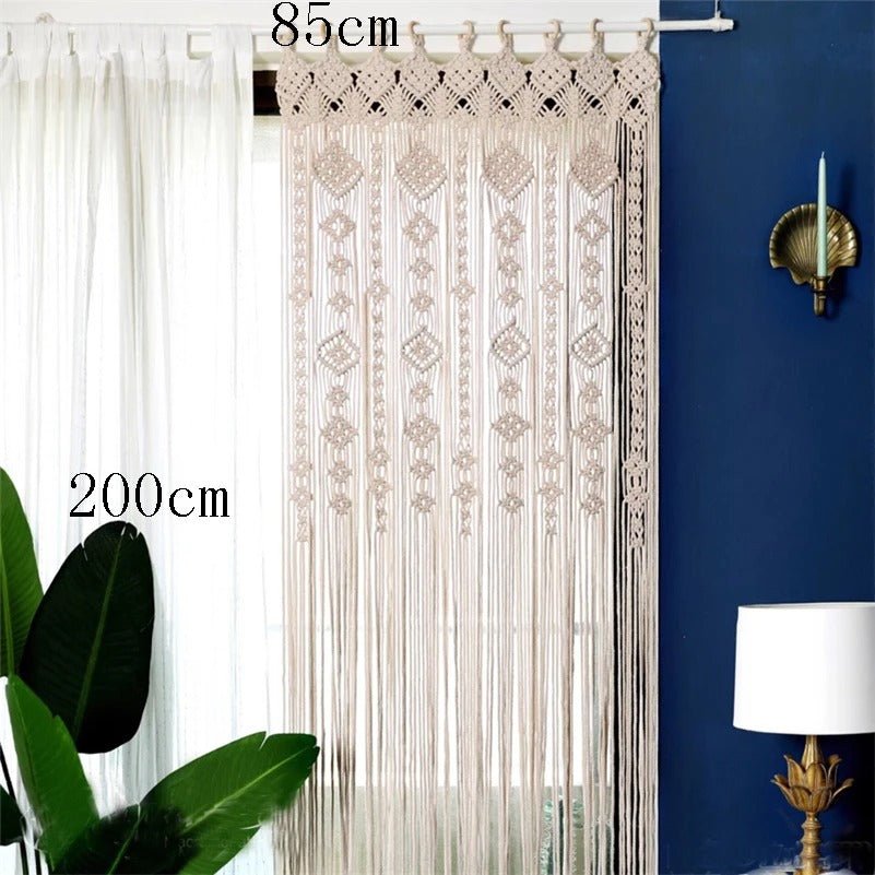 Un macramé rideau beige clair, mesurant 85 cm de large et 200 cm de haut, est suspendu à une tringle blanche devant une fenêtre partiellement cachée par un rideau blanc transparent. Le mur derrière est peint en bleu foncé et agrémenté d'une applique murale dorée en forme de coquillage tenant une bougie blanche. Au premier plan, à gauche, apparaissent de grandes feuilles vertes de plante.