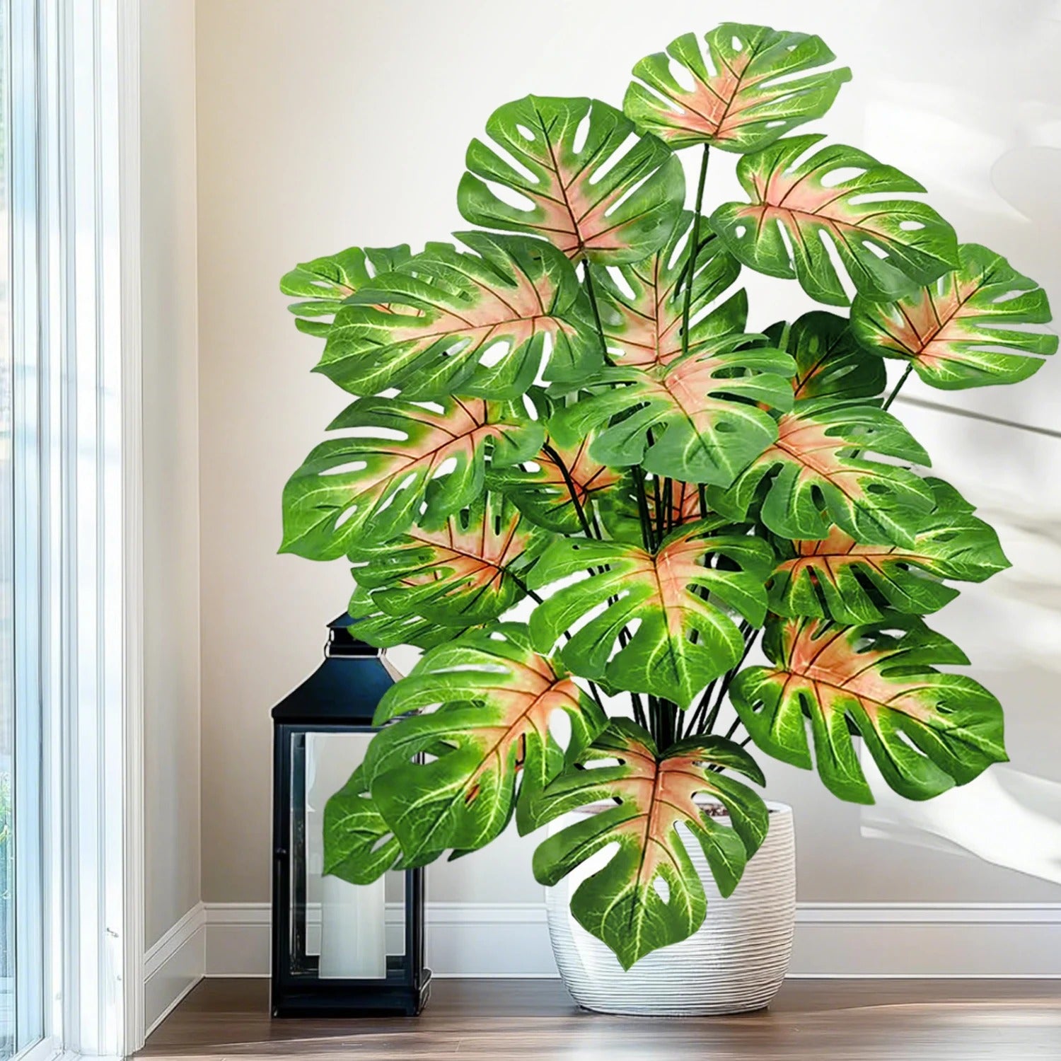 Un grand Plante Artificielle Monstera artificiel, aux feuilles vertes luxuriantes avec des panachures pêche, trône dans un pot blanc texturé.
La plante est placée près d'une fenêtre lumineuse, baignant la scène d'une douce lumière naturelle.
À gauche, une lanterne noire classique avec une bougie blanche ajoute une touche d'élégance à l'arrangement.
L'ensemble crée une ambiance intérieure chaleureuse et décorative sur un plancher en bois clair.