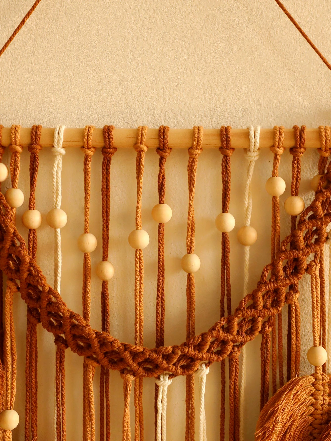 Un macramé décoratif aux tons chauds de brun et de beige est suspendu à un bâton de bois clair contre un mur crème. Des cordes nouées et des perles de bois forment un motif texturé et artisanal. La lumière douce met en valeur les détails du marcrame. L'ensemble crée une ambiance bohème et chaleureuse.