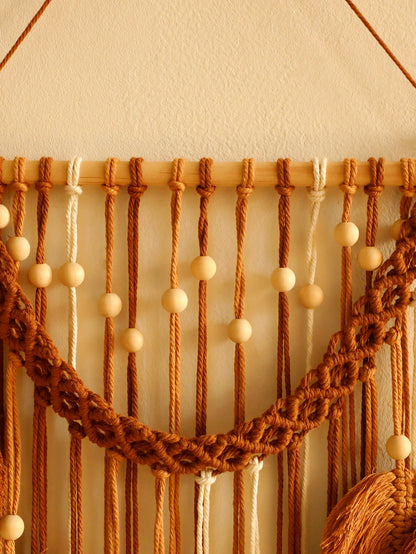 Un macramé décoratif aux tons chauds de brun et de beige est suspendu à un bâton de bois clair contre un mur crème. Des cordes nouées et des perles de bois forment un motif texturé et artisanal. La lumière douce met en valeur les détails du marcrame. L'ensemble crée une ambiance bohème et chaleureuse.