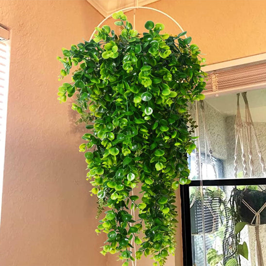 Cette image met en scène un panier suspendu blanc contenant de luxuriantes plantes artificielles retombantes. Les fausses feuilles vertes, denses et variées, créent un effet de cascade végétale décorative. L'ensemble est suspendu devant un mur de couleur pêche et une fenêtre partiellement visible. On aperçoit également des stores et un autre arrangement de plantes en macramé en arrière-plan. Ces plantes artificielles retombantes apportent une touche de verdure sans nécessiter d'entretien.