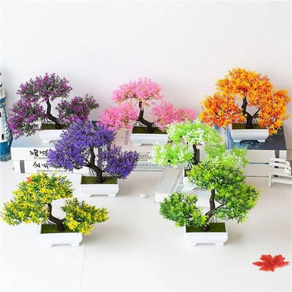 La photo présente un assortiment de petites plantes artificielles disposées sur une surface blanche. Ces plantes artificielles imitent des bonsaïs avec des feuillages de différentes couleurs vives : violet, rose, orange, vert clair et vert foncé. Chaque plante artificielle est placée dans un petit pot carré blanc. L'ensemble crée une décoration colorée et sans entretien grâce à ces plantes artificielles.