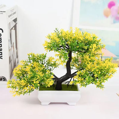 Sur un fond blanc épuré, on observe une plante artificielle luxuriante. Ses feuilles, d'un vert vif parsemé de touches jaunes, imitent un feuillage dense et printanier. Le tronc noir torsadé repose dans un pot carré blanc aux bords légèrement ondulés. Cette plante artificielle apporte une touche de verdure sans nécessiter d'entretien. Elle est placée près de livres et d'une image, suggérant un décor intérieur soigné.