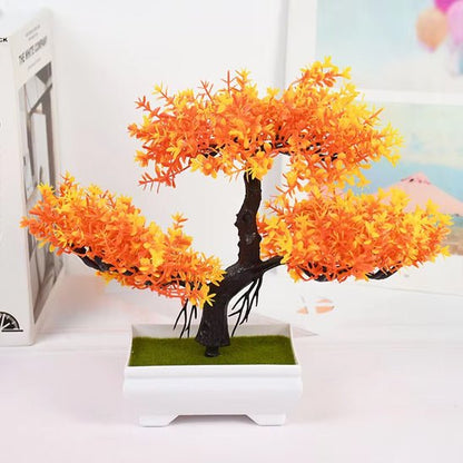 Cette image présente une plante artificielle de type bonsaï aux couleurs automnales, avec des feuilles orange et jaune vif. Elle est placée dans un pot carré blanc contenant une imitation de mousse verte. L'ensemble repose sur une surface blanche, avec un livre et un tableau flou en arrière-plan. La lumière met en valeur les détails de cette plante artificielle décorative.