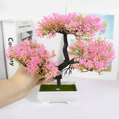 Sur cette image, on voit une main tenant une partie d'une plante artificielle ressemblant à un bonsaï en fleurs roses. La plante est disposée dans un petit pot carré blanc rempli d'une imitation de mousse verte. En arrière-plan, on aperçoit des livres et une carte floue. La lumière est douce et met en valeur les couleurs pastel de la plante artificielle. L'ensemble crée une atmosphère décorative et délicate.