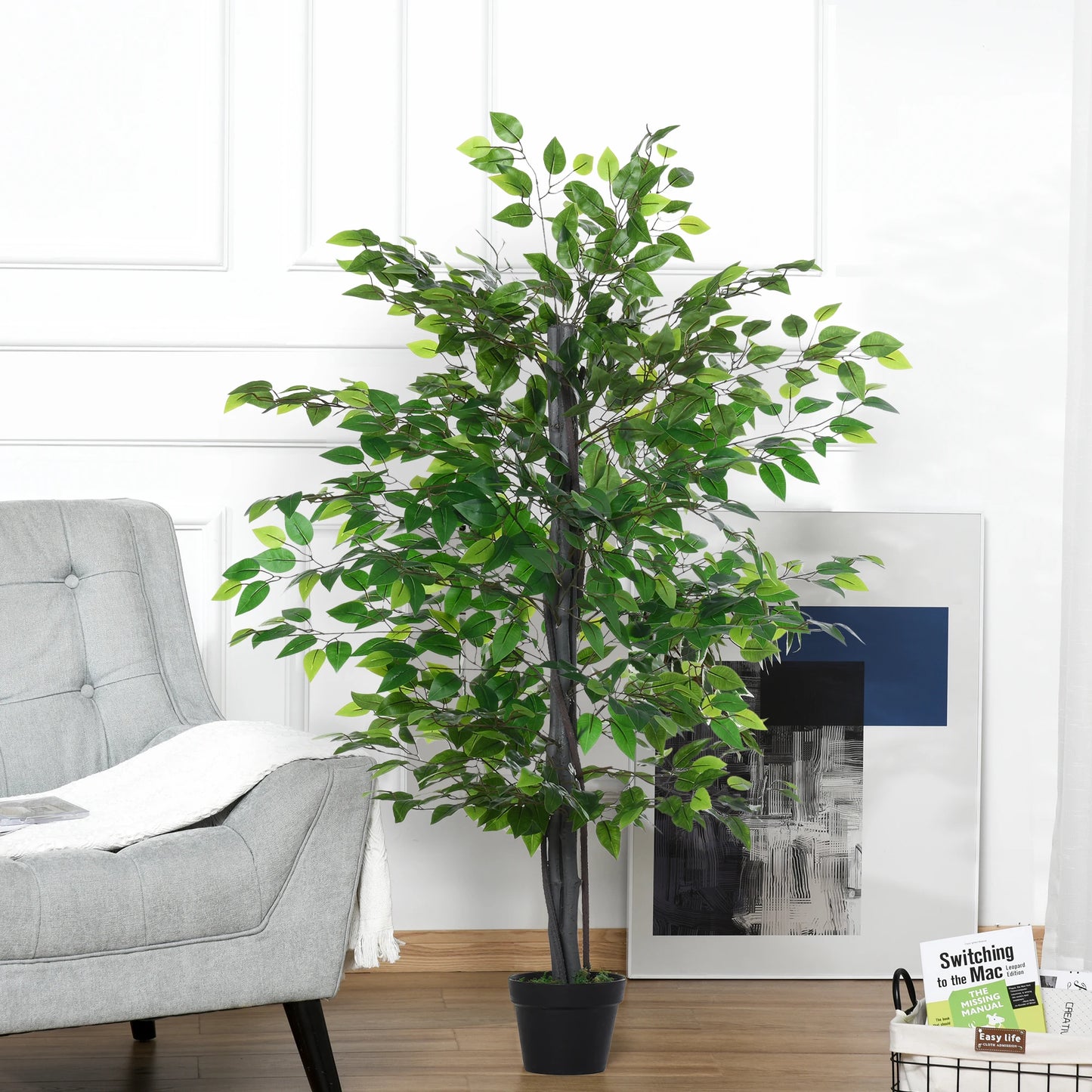 Une grande plante artificielle-Ficus aux feuilles vertes luxuriantes se dresse sur un plancher en bois à côté d'une chaise gris clair, contre un mur blanc avec des œuvres d'art abstraites encadrées et une petite pile de magazines en arrière-plan.