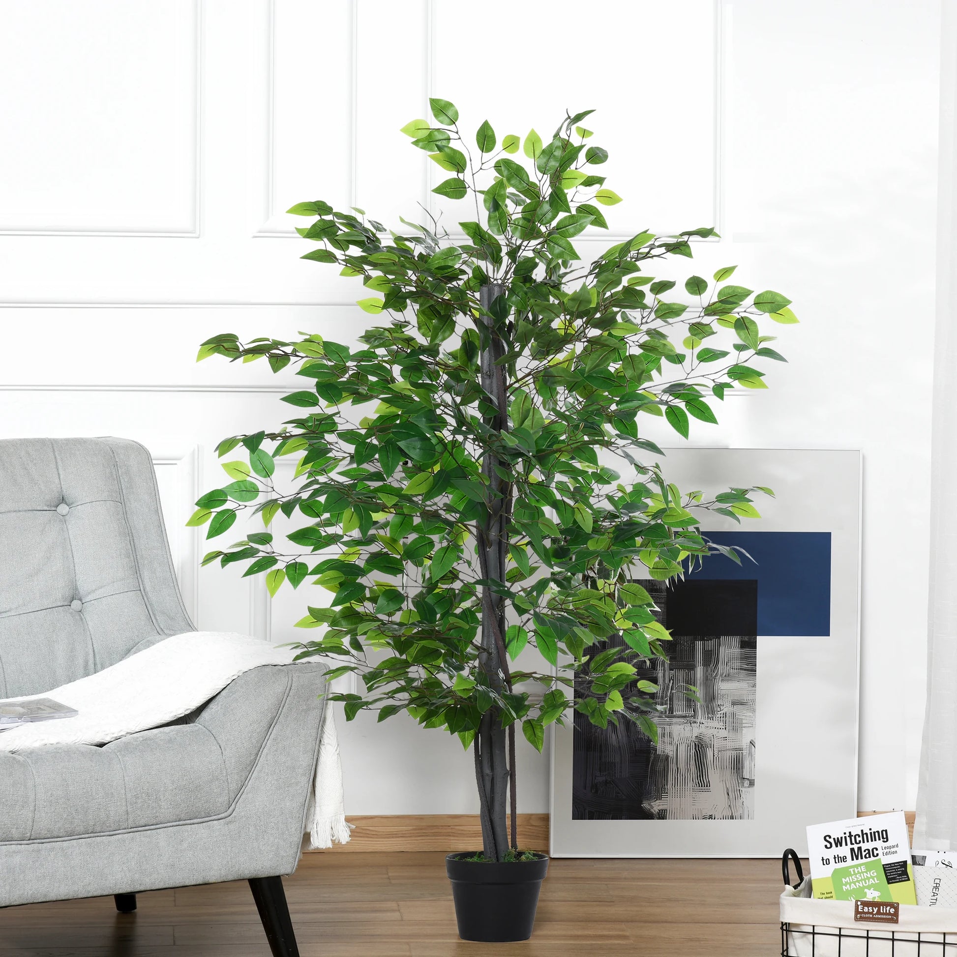 Une grande plante artificielle-Ficus aux feuilles vertes luxuriantes se dresse sur un plancher en bois à côté d'une chaise gris clair, contre un mur blanc avec des œuvres d'art abstraites encadrées et une petite pile de magazines en arrière-plan.