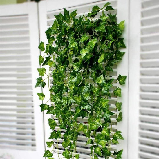 La photo présente un arrangement de plantes artificielles retombantes fixées sur un fond blanc à lamelles horizontales. Ces fausses lianes de lierre arborent des feuilles vertes aux nuances variées, créant un effet de cascade végétale. Elles sont disposées de manière à couvrir une partie significative du fond, apportant une touche de verdure artificielle à l'espace. La lumière naturelle ou artificielle met en évidence les détails des feuilles et leur texture. L'ensemble donne une impression de fraîcheur déc