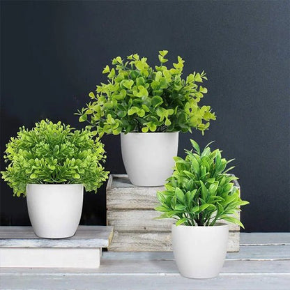 Trois plantes vertes artificielles Mini Plantes, idéales pour la décoration intérieure, sont disposées sur une surface gris clair sur un fond gris foncé. Dans de simples pots blancs – deux sur des caisses en bois empilées, un en surface – elles apportent une beauté naturelle sans aucun entretien.