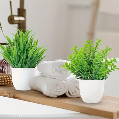 Deux petites plantes vertes en pot, peut-être des Mini Plantes (plante verte artificielle), sont posées sur un plateau en bois à côté de serviettes blanches roulées dans une salle de bain avec un robinet flou en arrière-plan, créant une atmosphère fraîche et apaisante qui incarne une décoration intérieure sans effort.