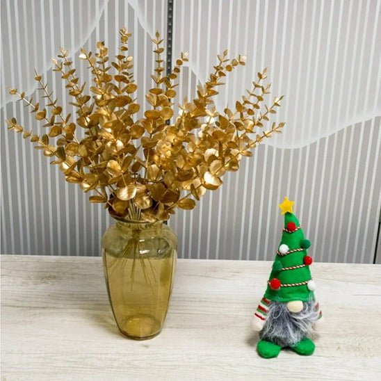 Un vase avec des Plantes artificielles comme des vraies-Feuilles D'eucalyptus Doré se dresse sur une surface en bois clair à côté d'une décoration d'arbre de Noël en peluche avec une étoile, des perles rouges et une fausse barbe grise - une charmante décoration d'intérieur.