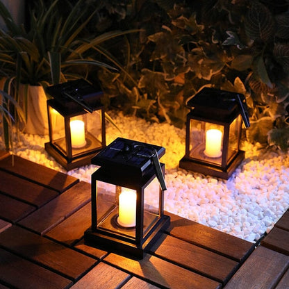 Trois lanternes Lampe solaire jardin haut de gamme-Classic-Solar™ brillent sur une terrasse en bois à côté de galets blancs et de plantes vertes, créant une ambiance extérieure chaleureuse et confortable la nuit avec un design d'inspiration rétro.